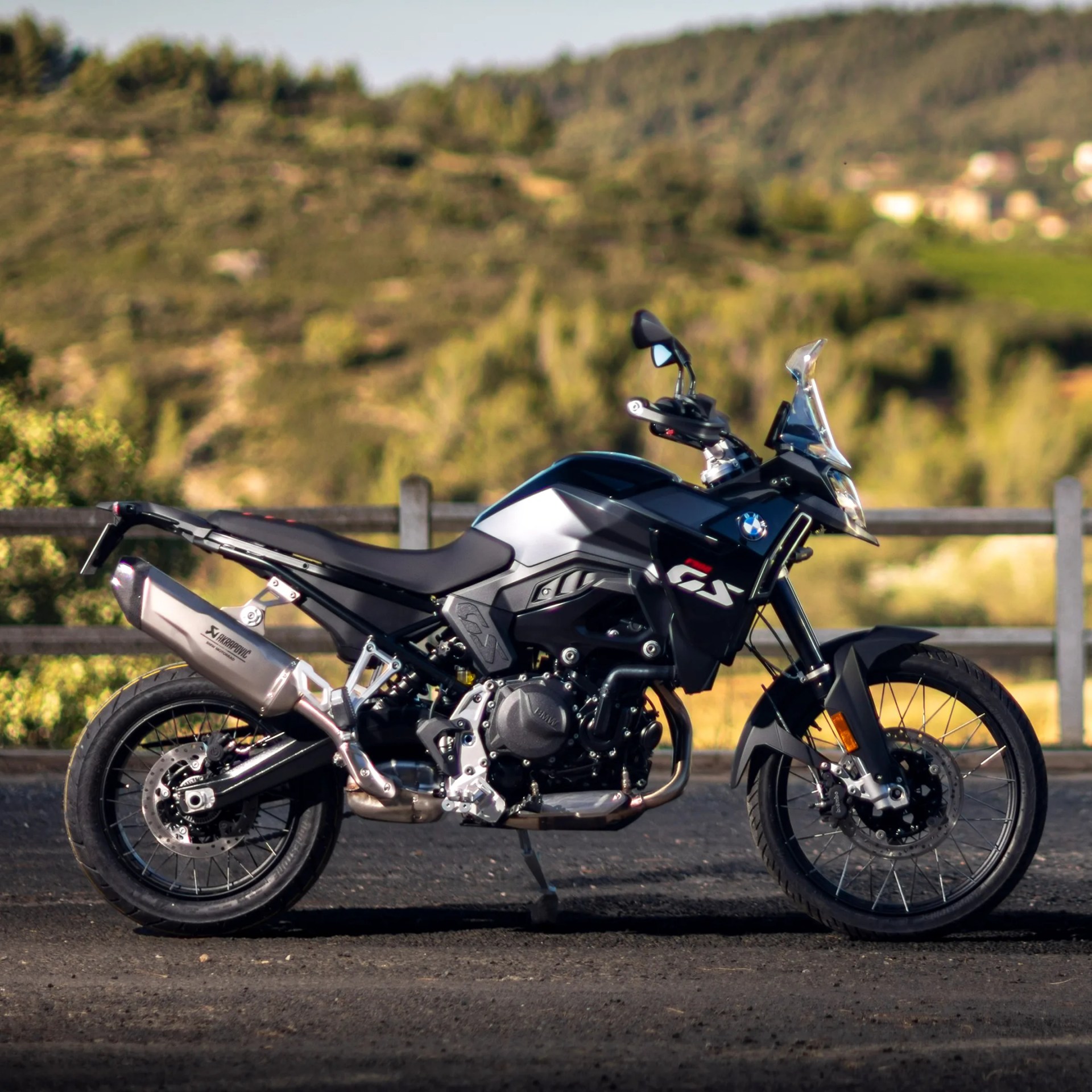 Black and silver BMW GS motorcycle parked on a paved surface with a wooden fence and green hills in the background. The bike features a prominent exhaust, spoked wheels, and a clear windshield.