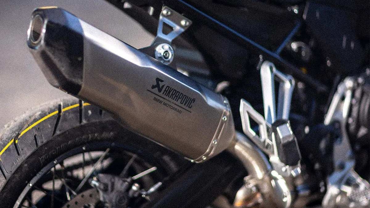 Close-up of a motorcycle's rear section featuring a silver Akrapovič exhaust system with "BMW Motorrad" branding, a black tire with a yellow stripe, and part of the footpeg assembly.