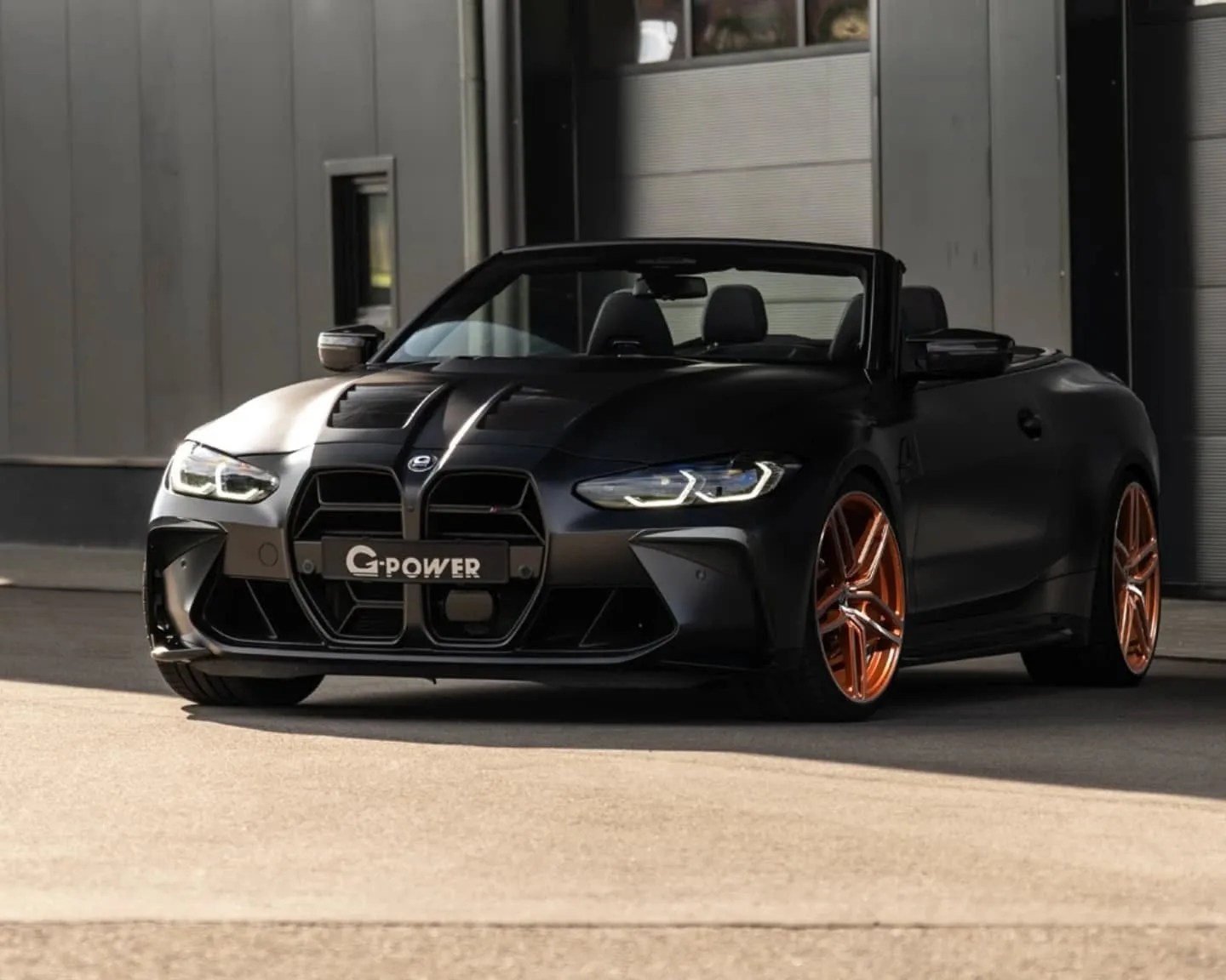 Black convertible sports car with a distinctive large front grille, sleek headlights, and bronze-colored alloy wheels, parked in front of a modern industrial building. The car has a "G-POWER" license plate.