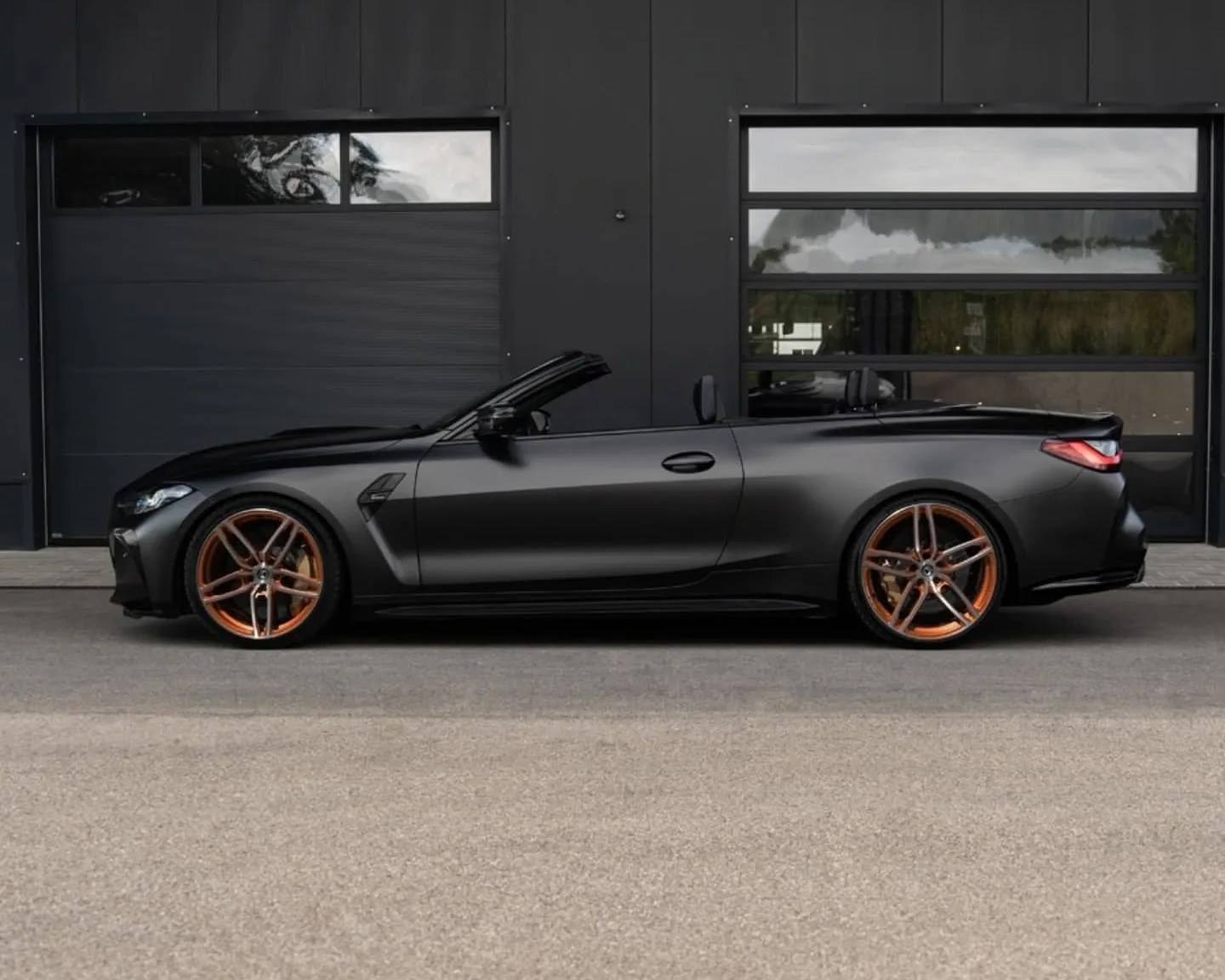 Matte black convertible sports car with copper-colored alloy wheels, parked in front of a modern black building with two large garage doors. The car's roof is down, showcasing its sleek, aerodynamic design.