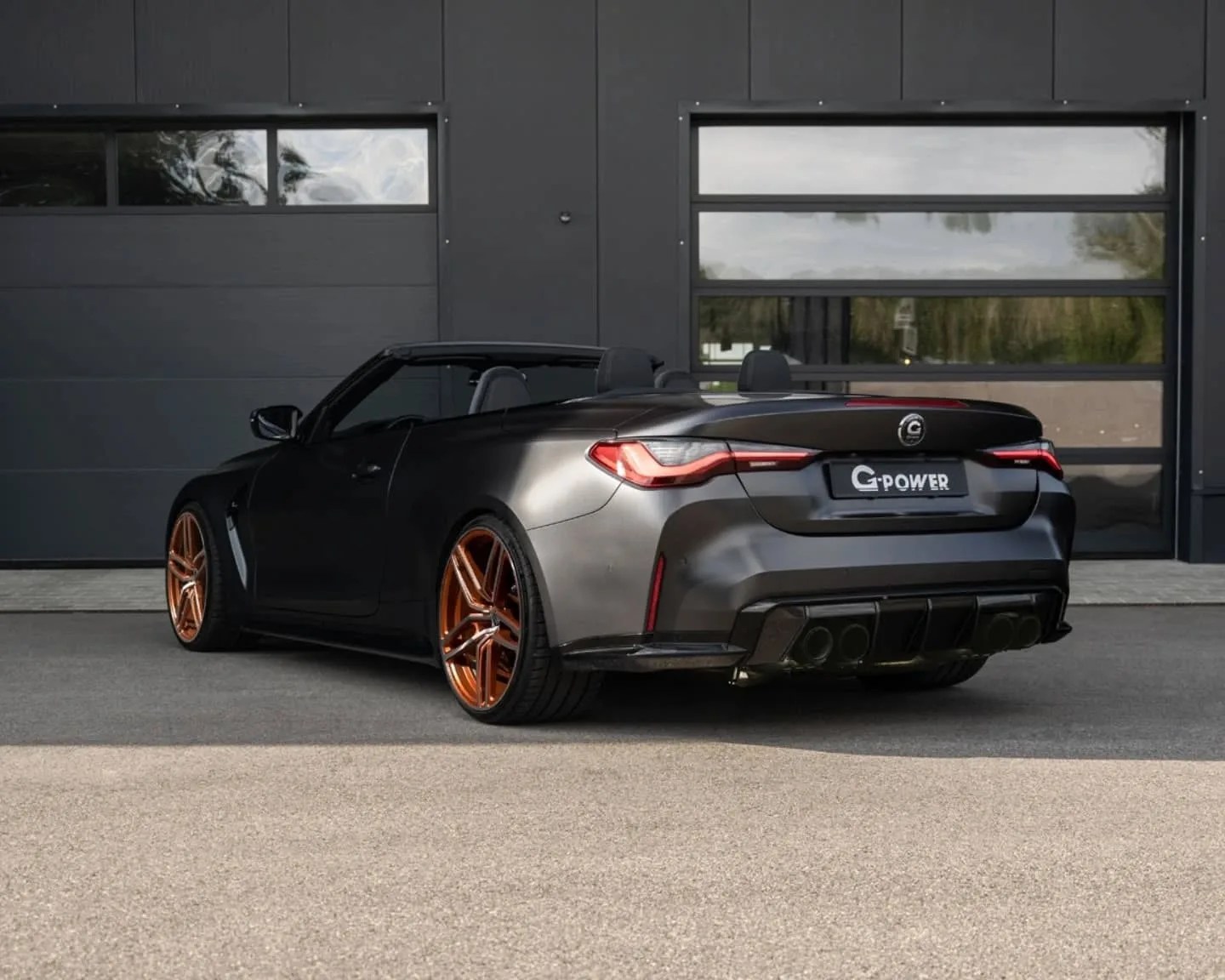 Black convertible sports car with a matte finish, featuring distinctive orange alloy wheels, quad exhaust pipes, and a "G-POWER" license plate, parked in front of a modern building with large dark garage doors.