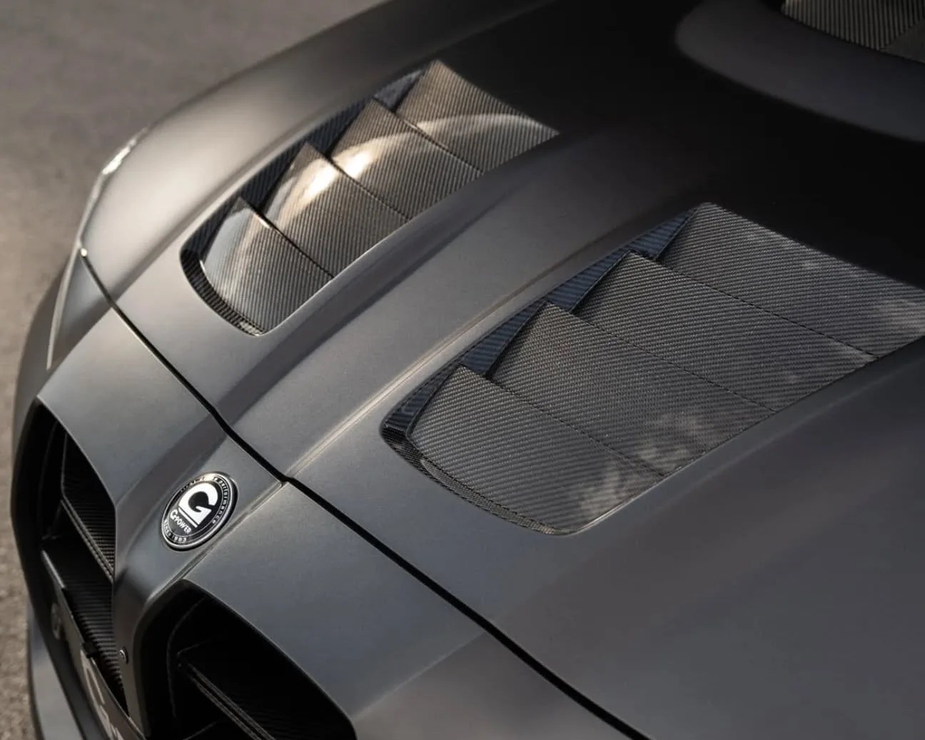 Close-up of a matte gray car hood featuring two carbon fiber vented sections and a circular emblem with the letter "G" and the word "Grotta" on it. The vents have a textured, layered design and reflect light subtly.