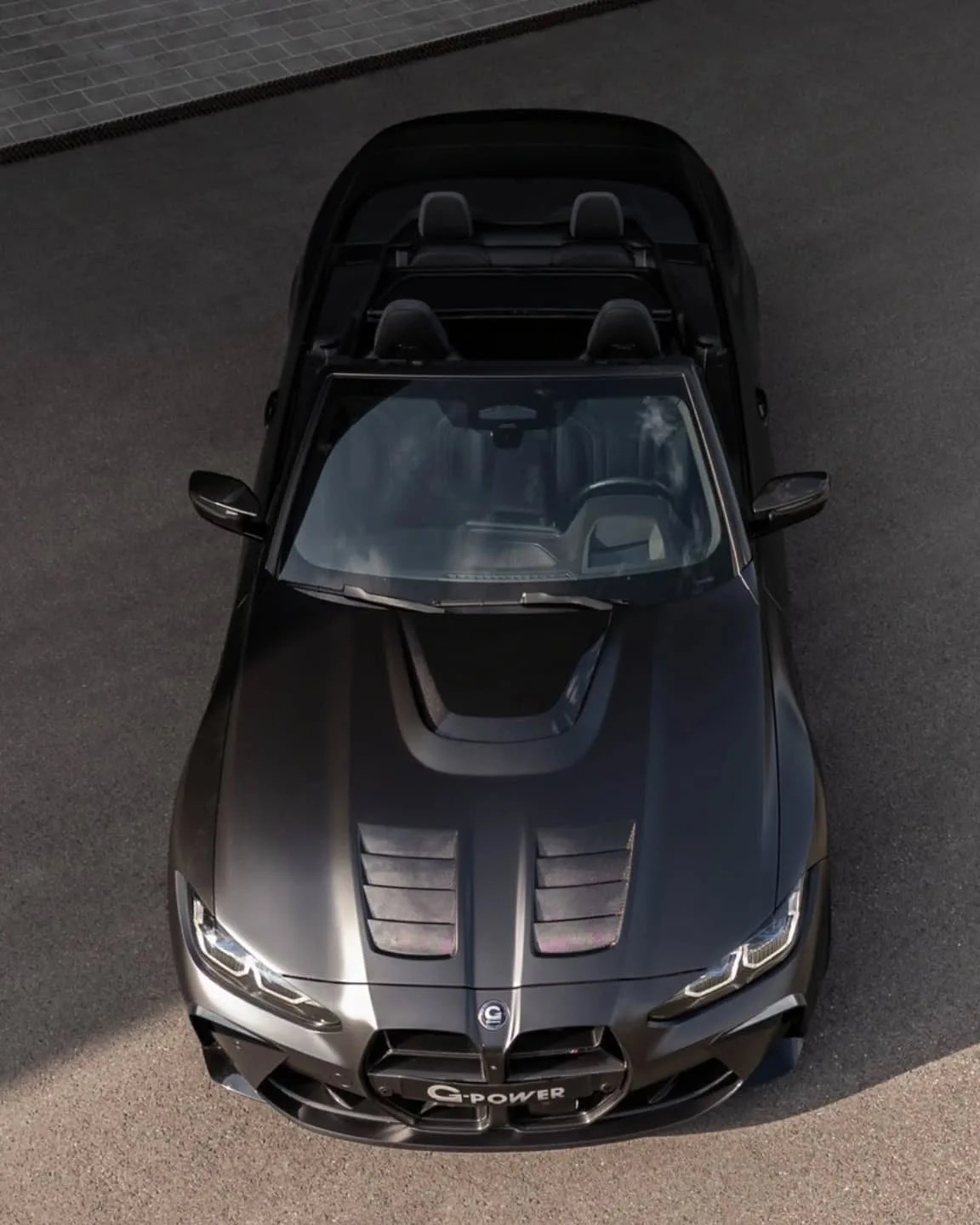Black convertible sports car with aggressive front design, featuring a prominent hood scoop and two vented sections on the hood. The car has sleek, angular headlights and a large front grille with a "G-POWER" badge. The interior shows four seats with headrests, and the car is parked on a paved surface.