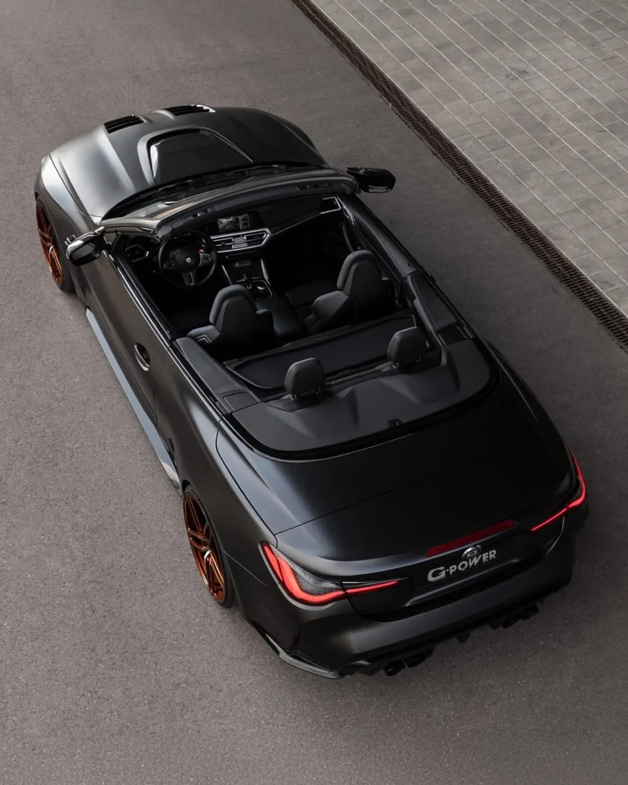 Matte black convertible sports car with the top down, featuring sleek aerodynamic lines, copper-colored alloy wheels, and modern LED tail lights. The interior is black with a digital dashboard and two front seats visible. The car is parked on a paved surface next to a tiled area with a drainage grate. The rear license plate reads "G-POWER.