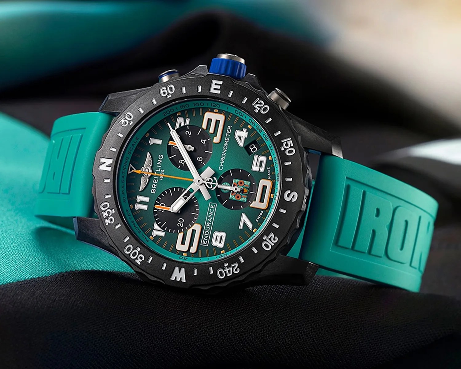 Breitling Endurance chronometer watch with a teal dial and black subdials, featuring large white and orange numerals and hands. The watch has a black bezel with white compass markings and a teal rubber strap embossed with the word "IRONMAN."
