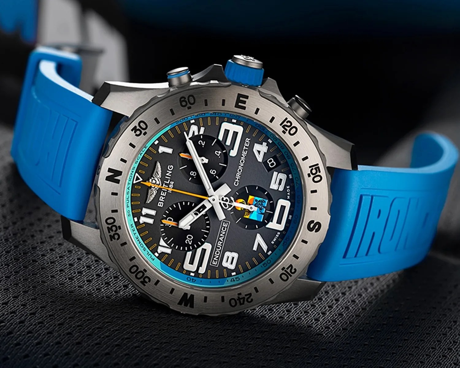 Breitling Endurance chronometer watch with a blue rubber strap, black dial featuring large white numerals, three subdials, and a date window, encased in a silver bezel marked with compass directions and minute indicators.