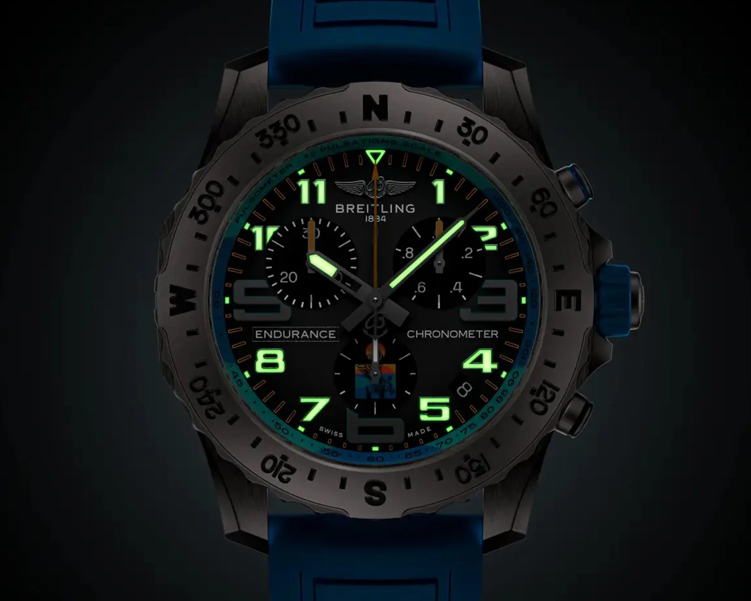 Breitling Endurance Chronometer watch with a black dial, luminescent green Arabic numerals and hands, three subdials, a blue rubber strap, and a black bezel marked with compass directions and degree increments.