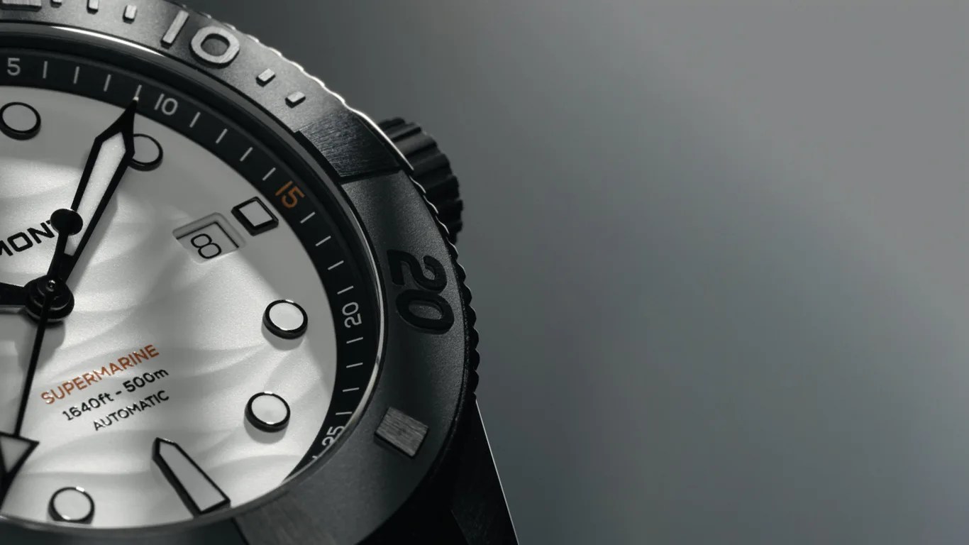 Close-up of a white and black bremont dive watch face showing the brand name partially, with the text "SUPERMARINE 1640ft - 500m AUTOMATIC" on the dial. The watch has a date window displaying the number 8, bold hour markers, and a rotating bezel with minute markings. The background is a smooth gradient of gray.