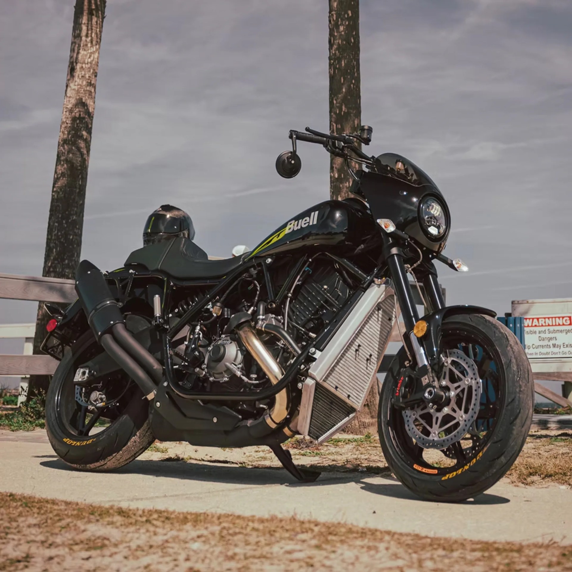 Black Buell motorcycle with a sleek design, featuring a round headlight, dual exhaust pipes, and Dunlop tires, parked on a concrete surface near palm trees and a wooden fence.