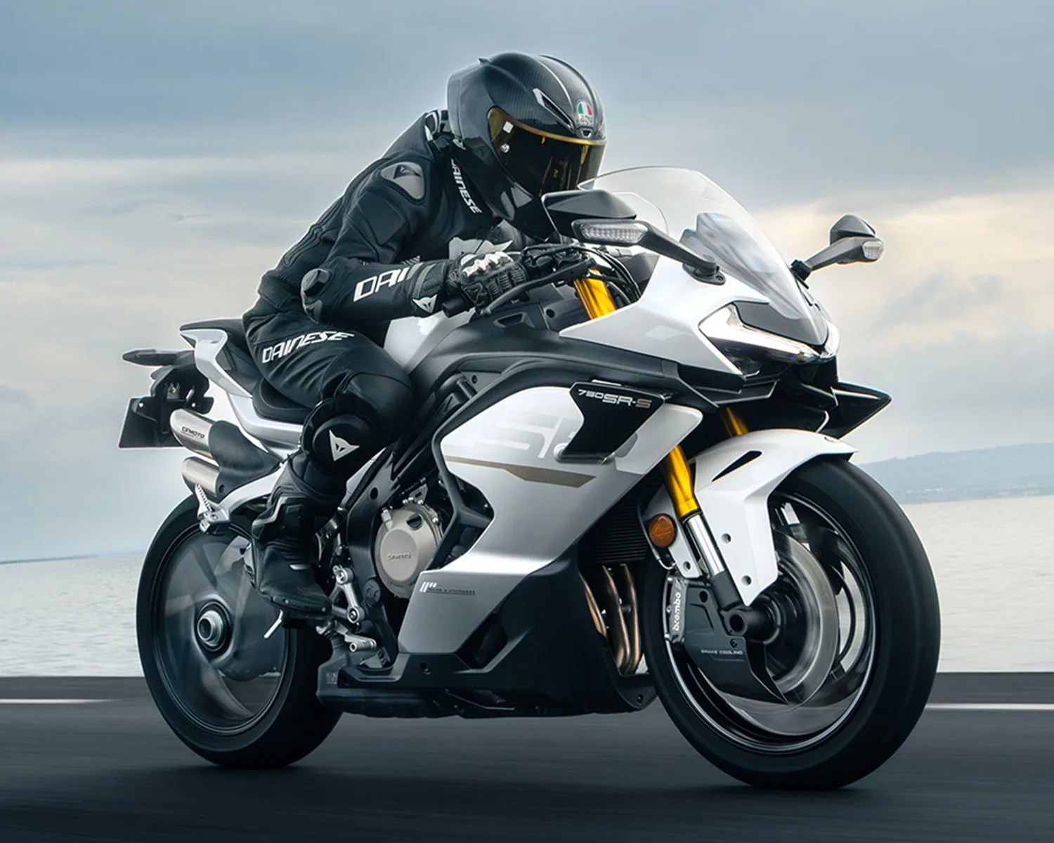 A rider in black protective gear and helmet is leaning forward on a white and black sport motorcycle with gold front forks, riding on a road near a body of water under a cloudy sky. The motorcycle has "780 SR-S" written on its side and features a sleek, aerodynamic design.