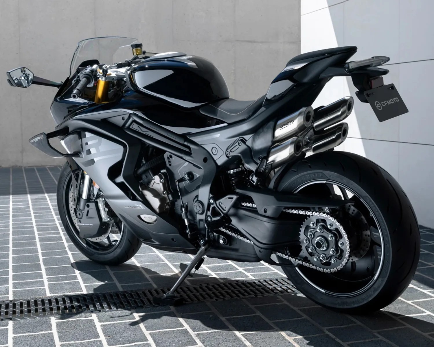 Black and silver sport motorcycle with a sleek, aerodynamic design, featuring dual exhaust pipes stacked on the right side, a clear windshield, and a visible chain drive on the rear wheel. The motorcycle is parked on a tiled surface against a concrete and white wall background.