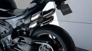 Rear section of a black motorcycle showing the rear tire, chain, and triple exhaust pipes with "CFMOTO" branding on the exhaust and license plate. The motorcycle has a sleek, modern design with a visible suspension spring and detailed mechanical components.