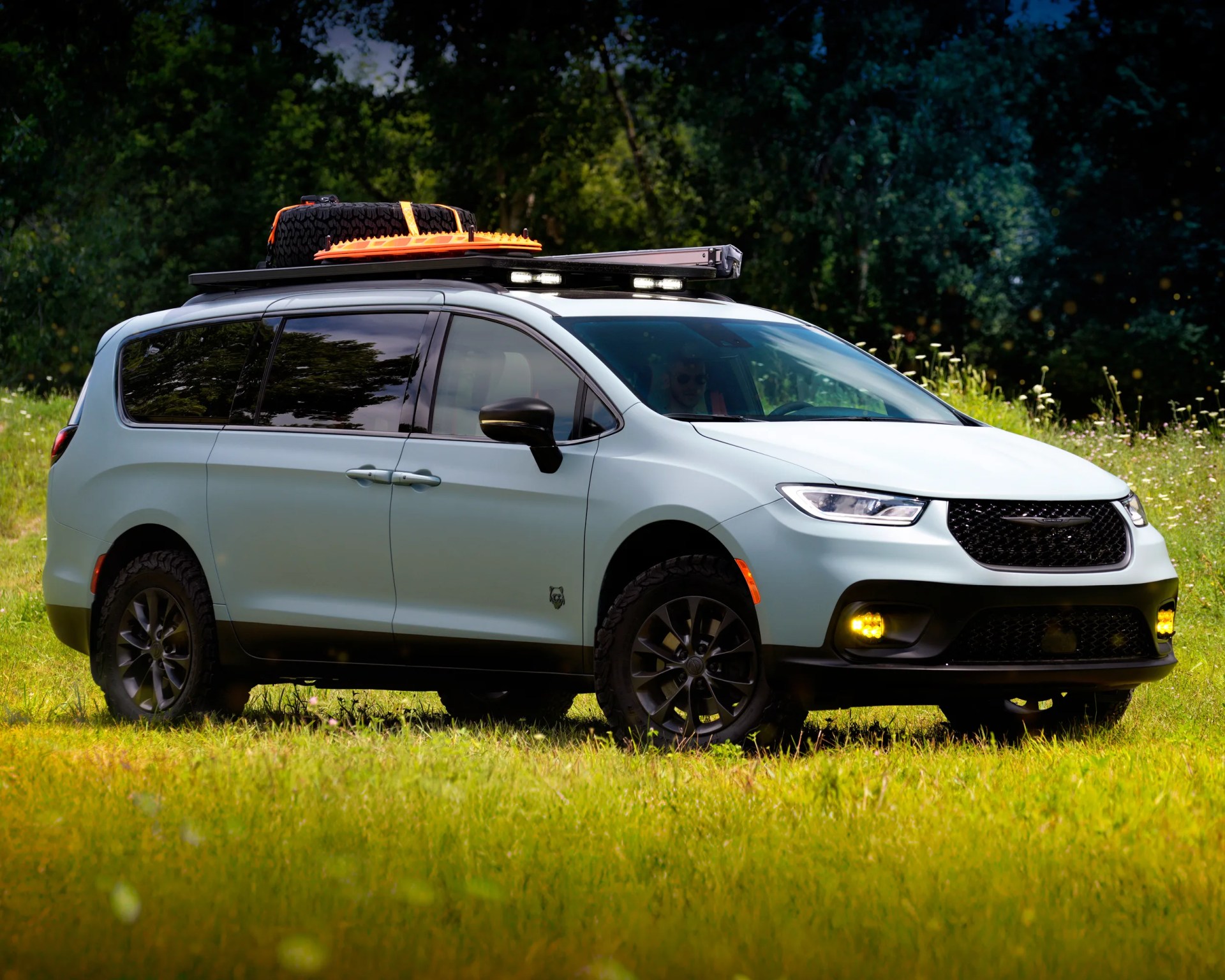 A light blue off-road minivan with black trim and rugged tires is parked on grass. It has a roof rack carrying a spare tire and orange recovery boards, along with mounted LED lights. The front fog lights are yellow, and the windows are tinted. The background features dense green trees.
