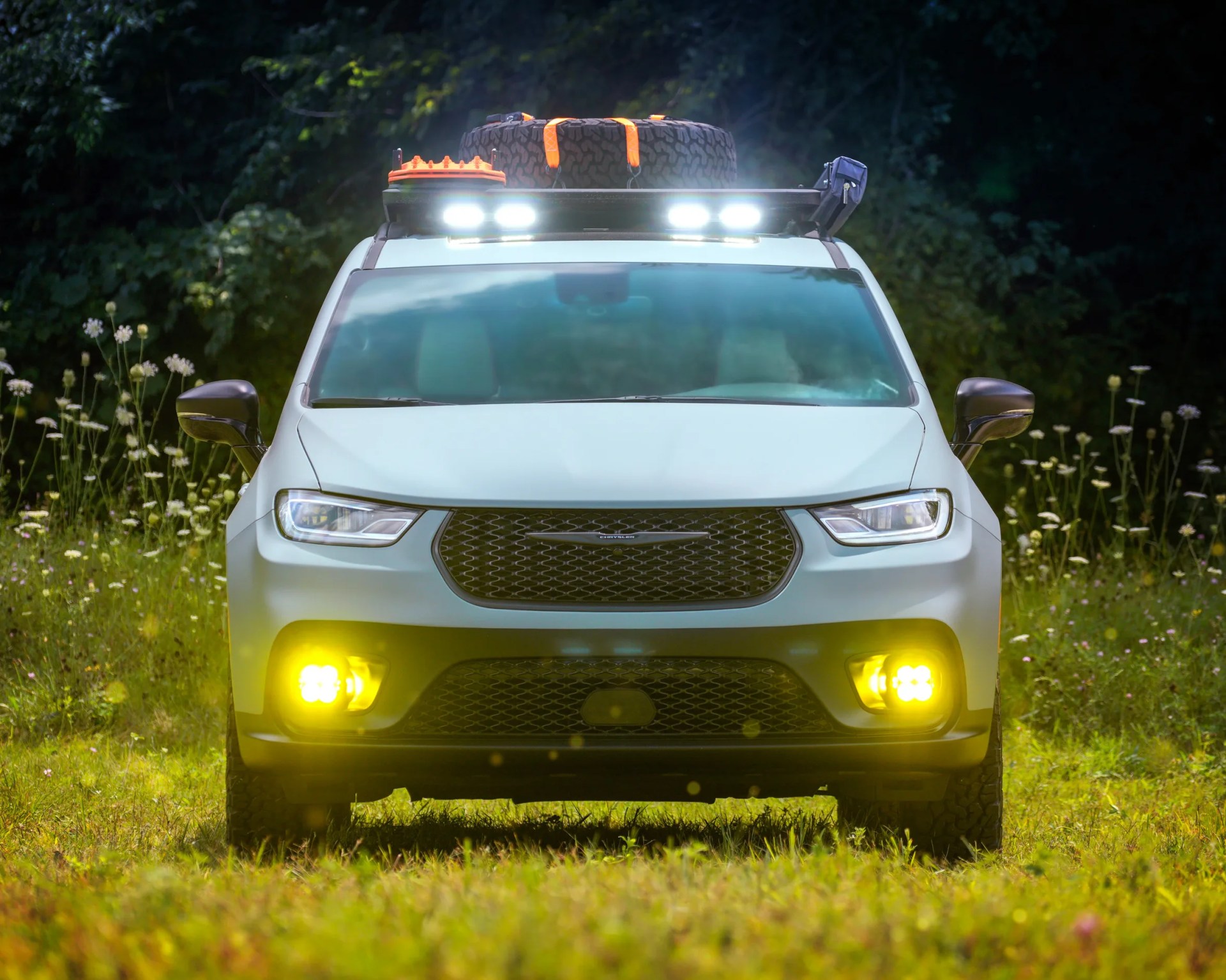 Front view of a white off-road vehicle with bright white roof lights and yellow fog lights turned on, parked on grass with wildflowers and dense trees in the background. The vehicle has a roof rack carrying two spare tires secured with orange straps and an orange container.