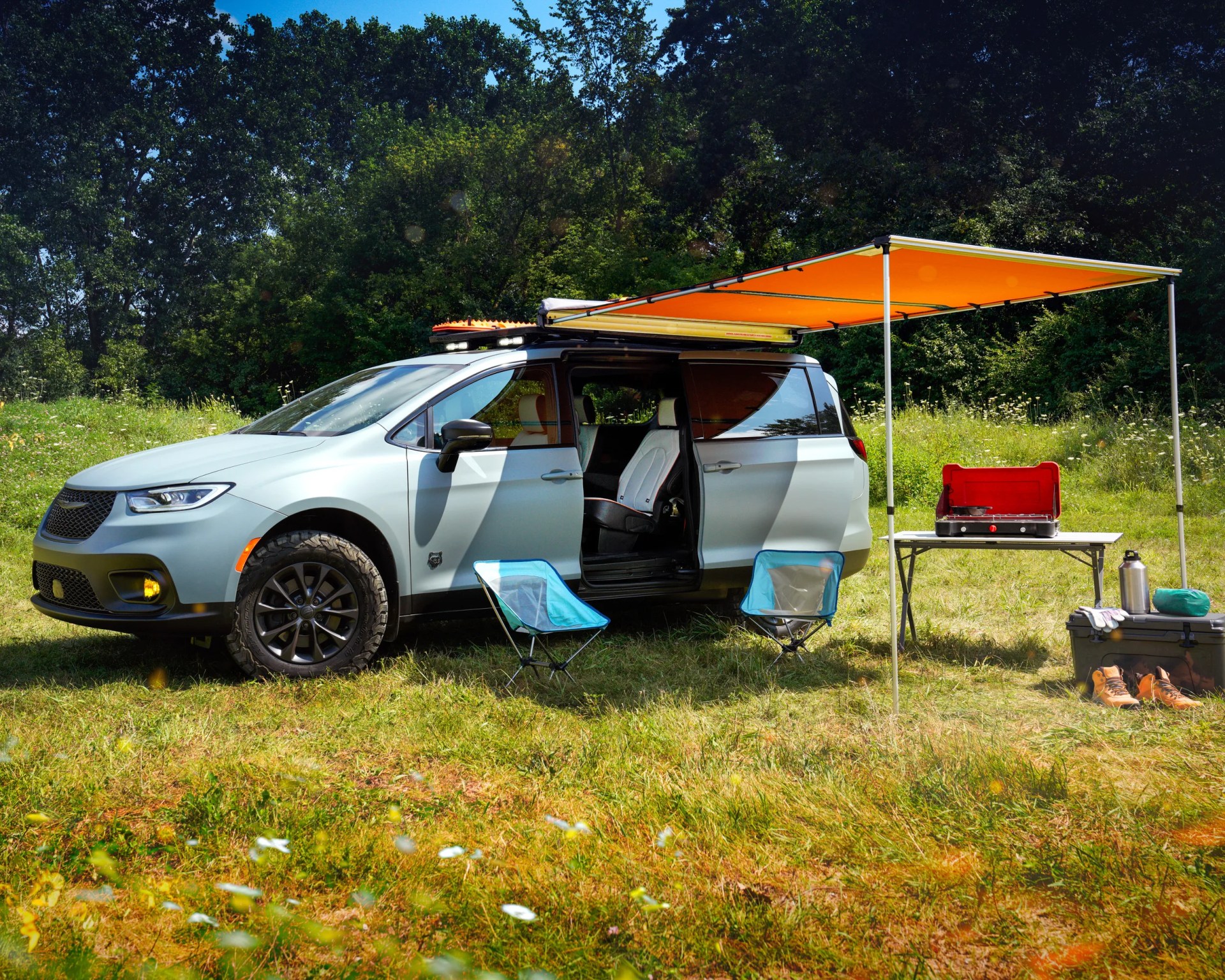 A light gray camper van is parked on a grassy field with its sliding door open. An orange awning extends from the side of the van, providing shade over two small blue camping chairs and a folding table. On the table, there is a red portable stove. Nearby, a gray storage box holds a pair of brown hiking boots, a water bottle, gloves, and a green bag. The background features dense green trees under a clear blue sky.