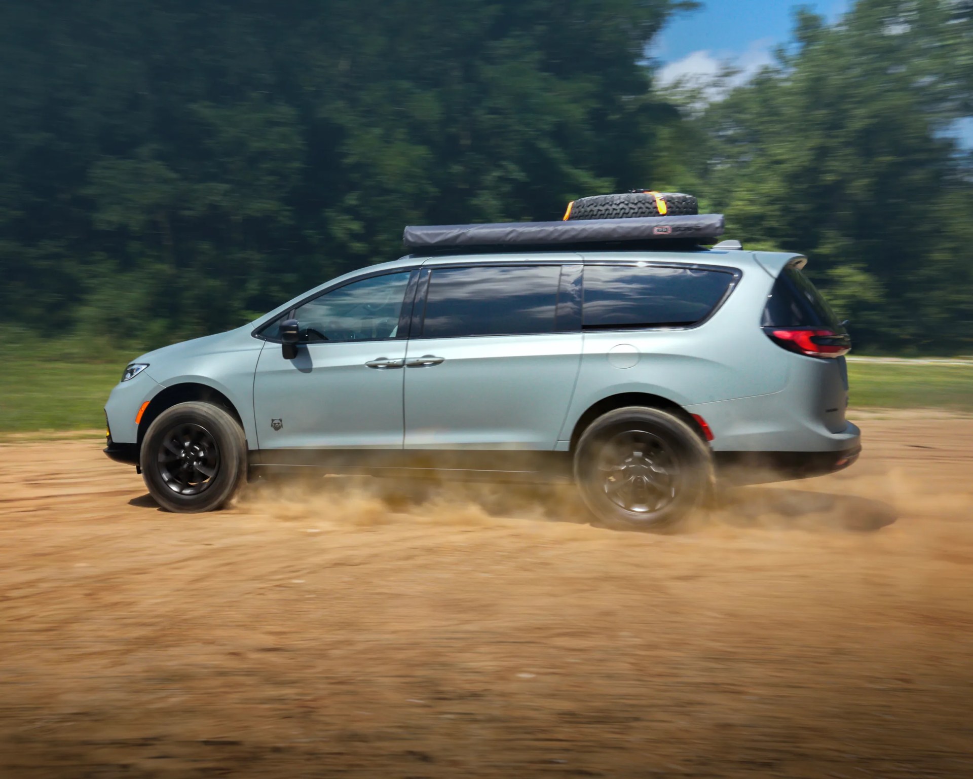 A light blue minivan driving on a dirt road, kicking up dust. The vehicle has black wheels, tinted windows, and a spare tire secured on the roof along with a rolled-up item, possibly a tent or awning. The background features green trees and a partly cloudy sky.