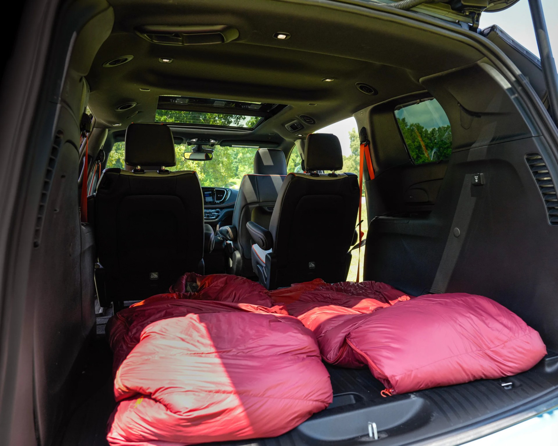 The interior of an SUV with the rear seats folded down, creating a flat space covered by two red sleeping bags. The front seats are upright, and the vehicle has a sunroof allowing natural light inside. The setting outside appears to be a green, wooded area.