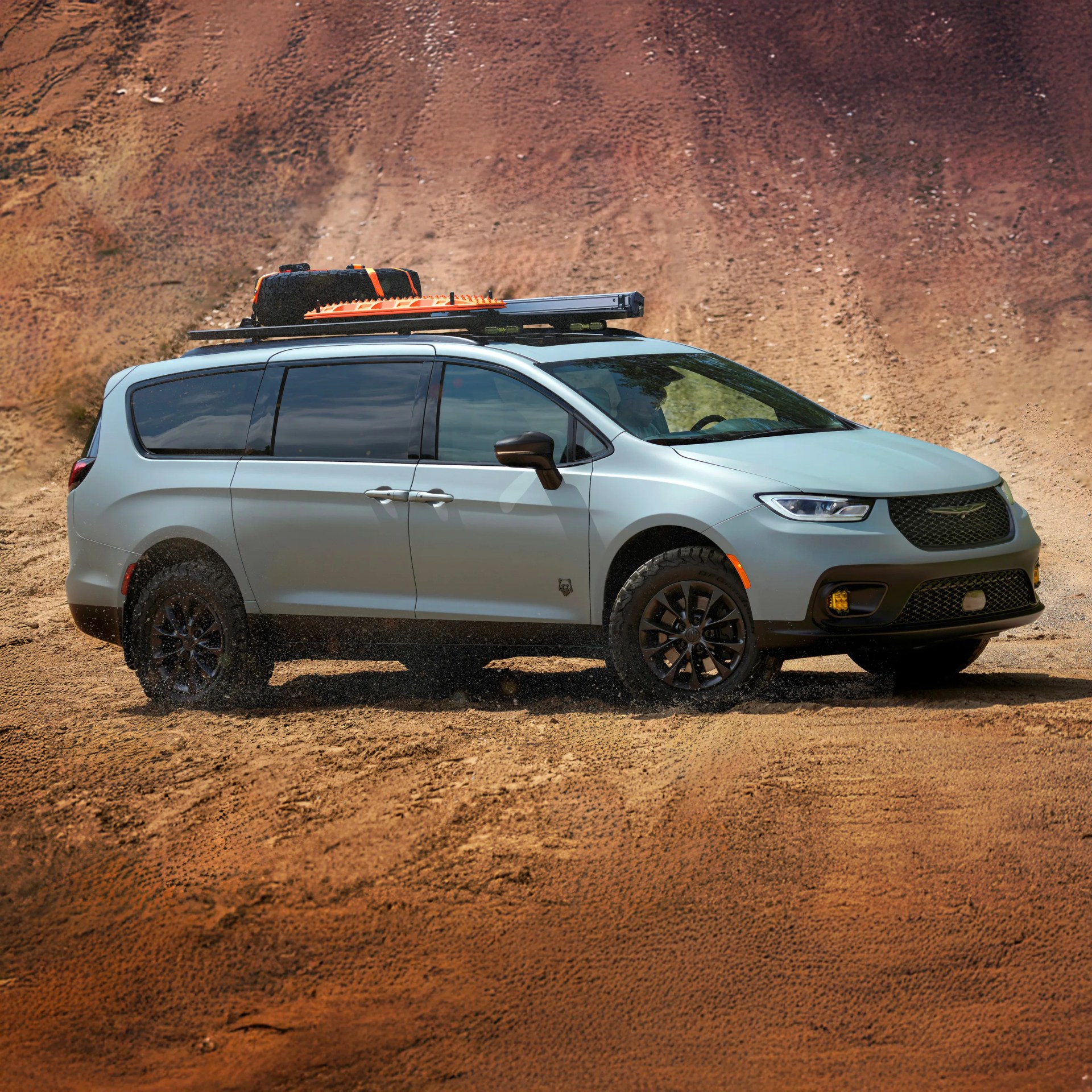 Silver-gray off-road minivan with black wheels and roof rack carrying a spare tire and orange recovery boards, parked on a sandy terrain with a reddish-brown dirt background.