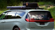 Light green Chrysler Pacifica minivan with a roof rack carrying a spare off-road tire secured with orange straps and a gray ARB Touring awning, parked outdoors with greenery in the background.
