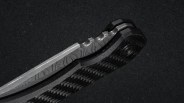 Close-up of a folding knife handle and partially visible blade. The blade has a patterned texture, likely Damascus steel, and the handle features a carbon fiber design with a textured grip and a pivot screw. The background is dark and textured.