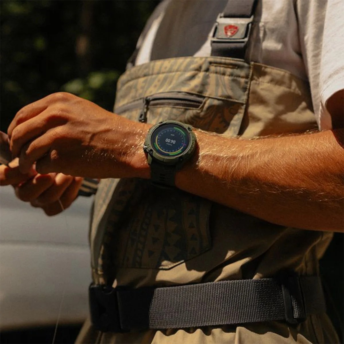 A person wearing a digital sports watch with a black strap on their left wrist. The watch face displays a timer or stopwatch with colorful circular progress indicators. The person is dressed in a beige patterned vest with a black buckle and a white shirt underneath. The background is outdoors with blurred greenery.
