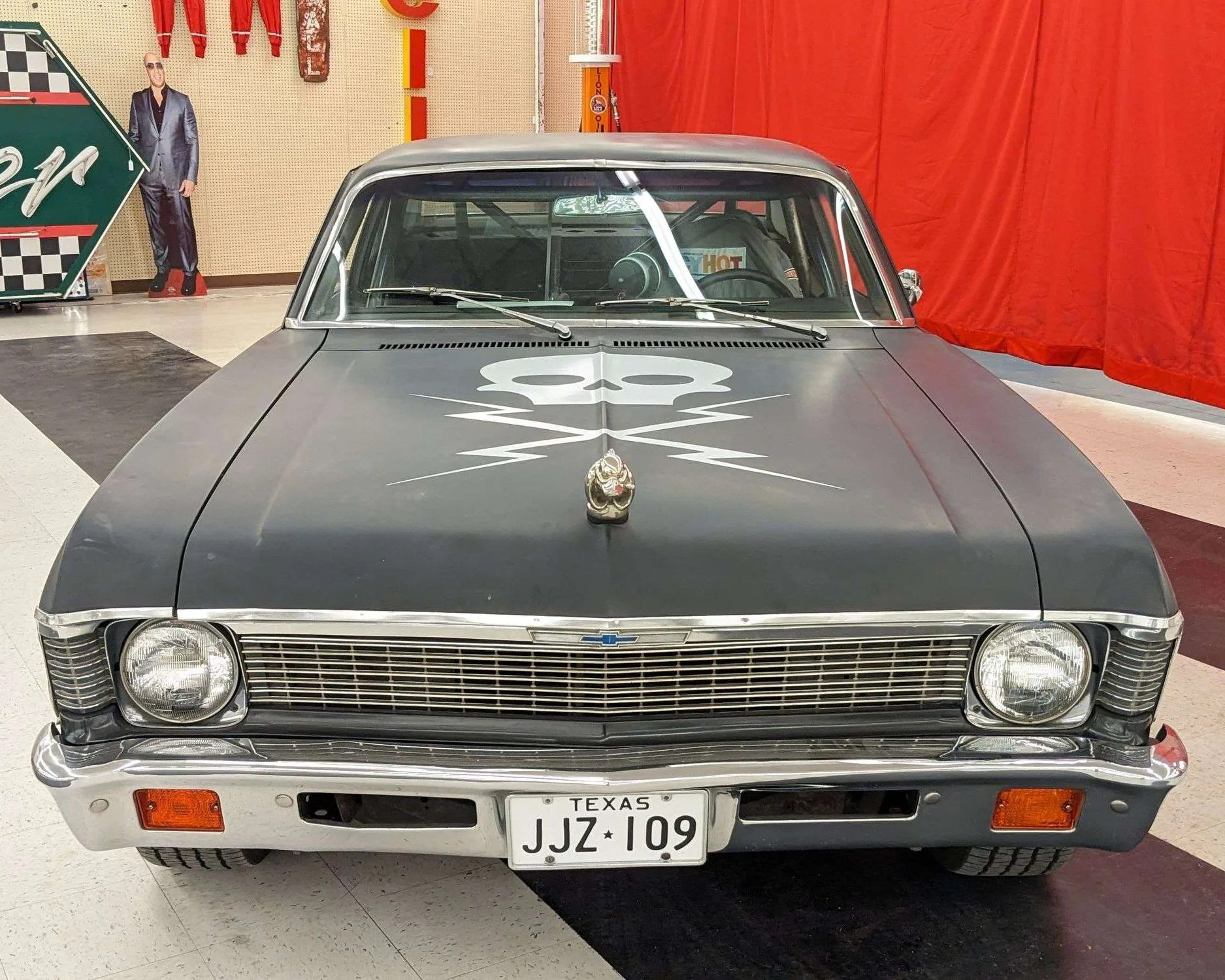 Front view of a matte black classic Chevrolet car with a skull and lightning bolt graphic on the hood, chrome grille and bumper, Texas license plate JJZ 109, and a hood ornament shaped like a clenched fist. The car is indoors with a red curtain in the background.