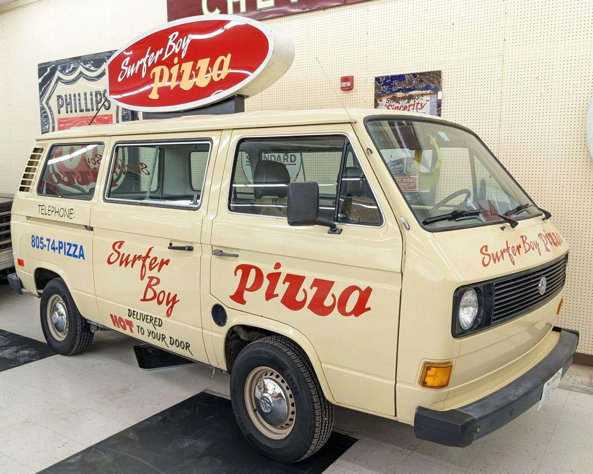 A beige Volkswagen van with red and blue lettering advertising "Surfer Boy Pizza." The van has a large red and white oval sign on its roof that reads "Surfer Boy Pizza." The side of the van displays the phone number "805-74-PIZZA" and the phrases "Surfer Boy" and "DELIVERED HOT TO YOUR DOOR." The front of the van also has "Surfer Boy PIZZA" written in red above the grille. The van is parked indoors on a tiled floor with vintage signs visible in the background.