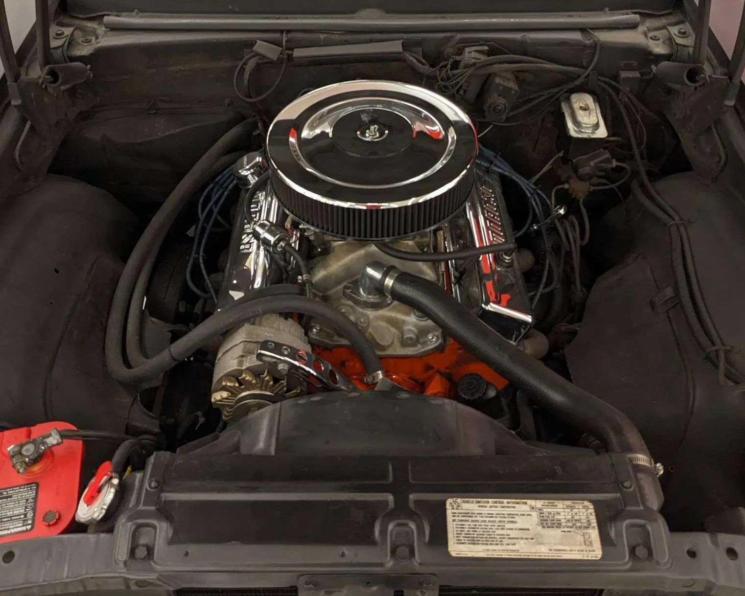 Car engine bay featuring a V8 engine with a large round chrome air filter cover on top, chrome valve covers, and visible black hoses. The engine block is painted orange, and there is a red battery located on the left side. The surrounding area is mostly black with some wiring and components visible.