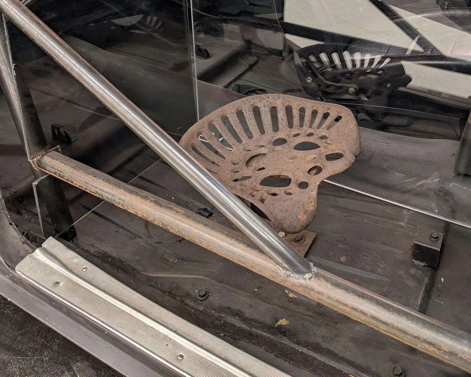 Rusty metal tractor-style seat mounted on the floor of a vehicle with a black interior and a metal roll cage bar running diagonally across the image. The seat has multiple cut-out holes and is reflected in a glass or clear plastic panel behind it.