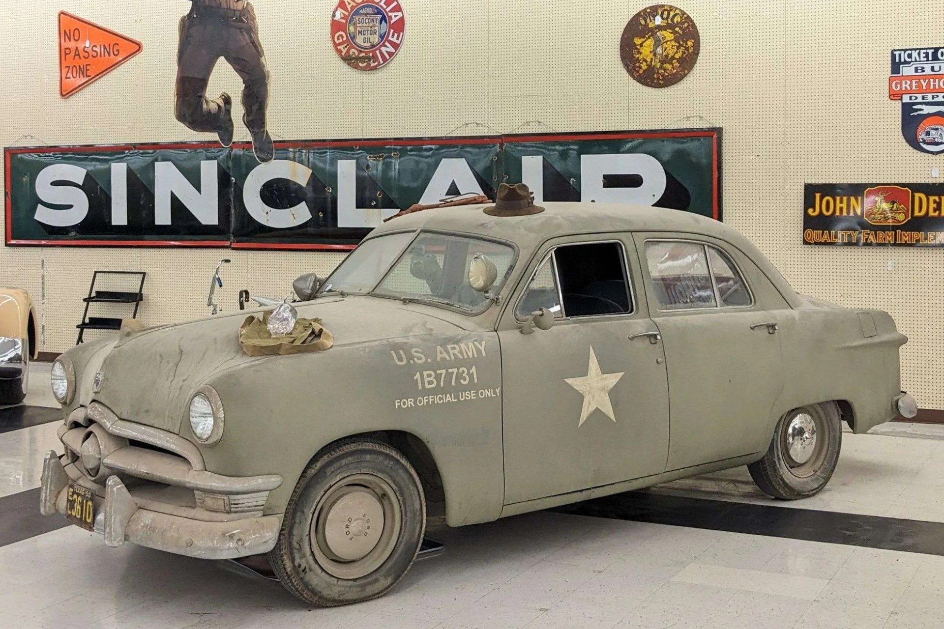 A vintage U.S. Army car painted in olive drab with a white star on the door and the text "U.S. ARMY 1B7731 FOR OFFICIAL USE ONLY" on the front fender. The car has a rounded body style typical of mid-20th century vehicles, with a flat tire on the front left wheel. A brown hat is placed on the roof, and there is a cloth with an object on the hood. The background features vintage signs, including a large "SINCLAIR" gasoline sign and other retro advertisements.
