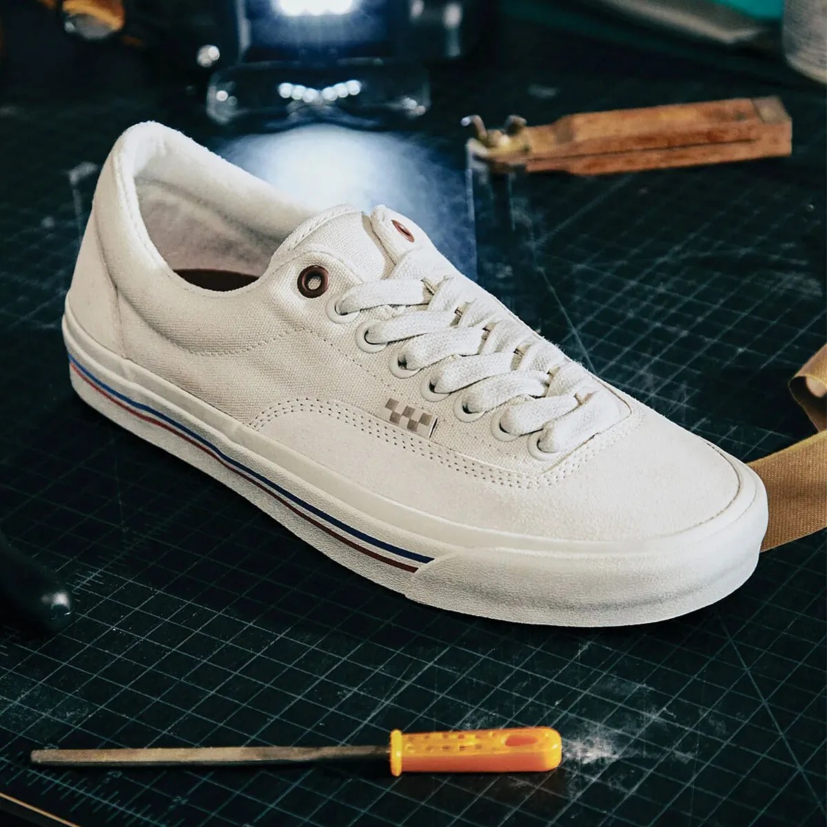 White canvas sneaker with white laces and a rubber sole featuring red and blue stripes, placed on a black grid-patterned surface with a yellow-handled screwdriver nearby.