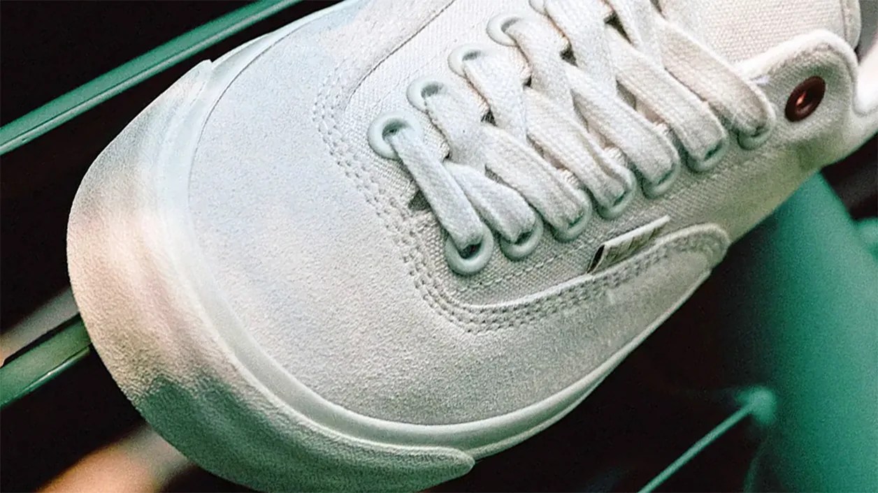 Close-up of a white sneaker with white laces, metal eyelets, and a textured rubber sole. The shoe appears to be made of canvas and suede materials.
