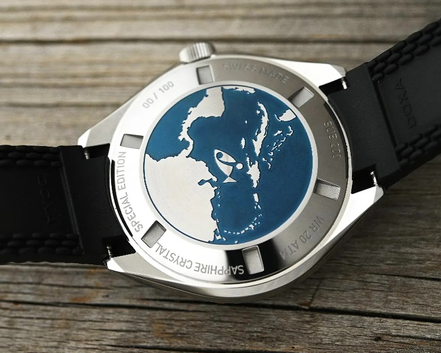 Doxa watch back case with a blue and white map design in the center, surrounded by engraved text including "SPECIAL EDITION," "SAPPHIRE CRYSTAL," "SWISS MADE," "SUB 200," and "WR 20 ATM." The watch has a black textured strap and is placed on a wooden surface.