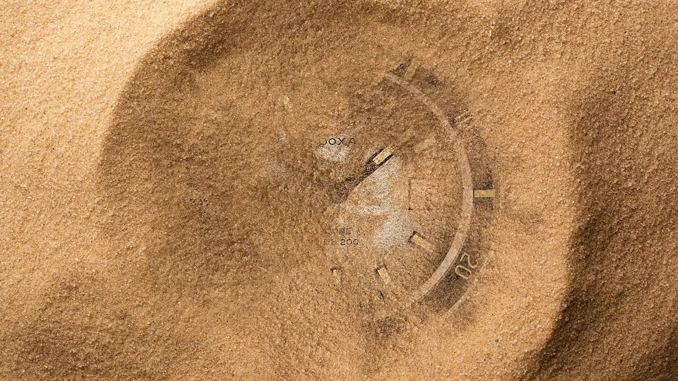 A partially buried doxa wristwatch in fine, light brown sand. The watch face is visible through the sand, showing a round dial with hour markers and a bezel marked with numbers, including "20". The watch hands are also faintly visible.