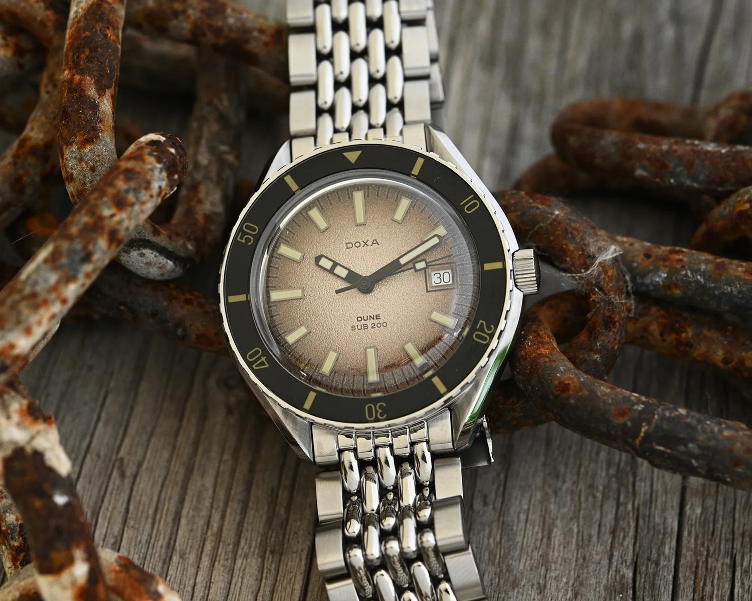 A close-up of a DOXA Dune Sub 200 wristwatch with a textured beige dial, luminous rectangular hour markers, and a date window at 3 o'clock. The watch has a black bezel with yellow markings and a stainless steel bracelet. It is placed on a weathered wooden surface surrounded by rusty chains.