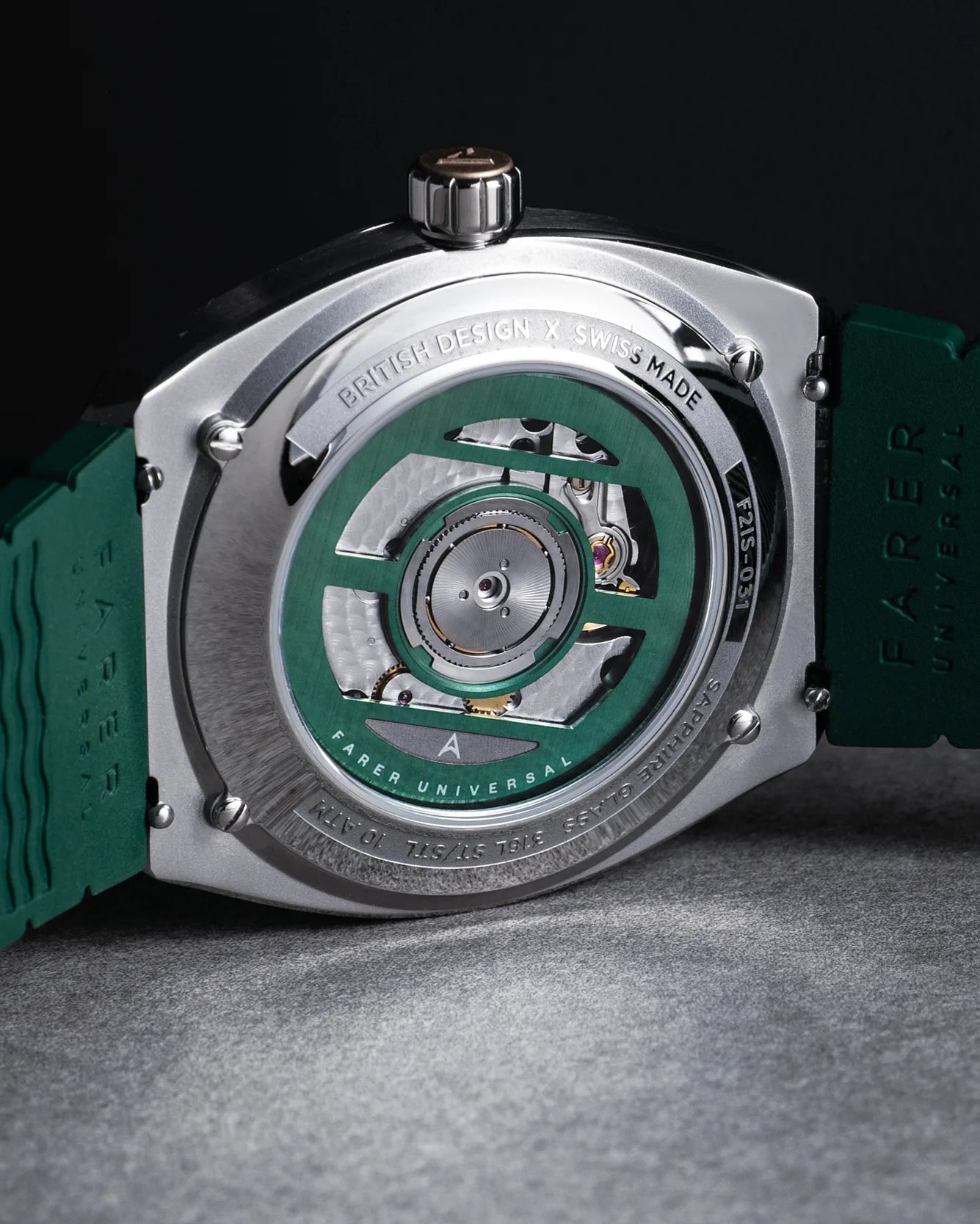 Close-up of the back of a Farer Universal wristwatch, showcasing its mechanical movement through a transparent sapphire glass case back. The watch features a green rotor with "FARER UNIVERSAL" engraved on it, a stainless steel case, and a green rubber strap. The case back is engraved with "BRITISH DESIGN X SWISS MADE," "SAPPHIRE GLASS," and other specifications. The crown is positioned at the top of the image.