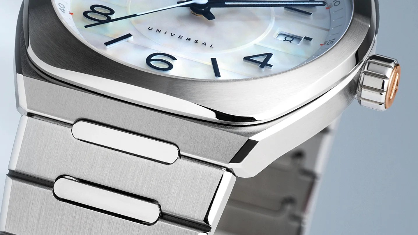 Close-up of a stainless steel wristwatch with a brushed metal bracelet and a polished bezel. The watch face features black numerals and hands, a date window, and the word "UNIVERSAL" printed on a mother-of-pearl dial. The crown has a two-tone design with a rose gold accent.