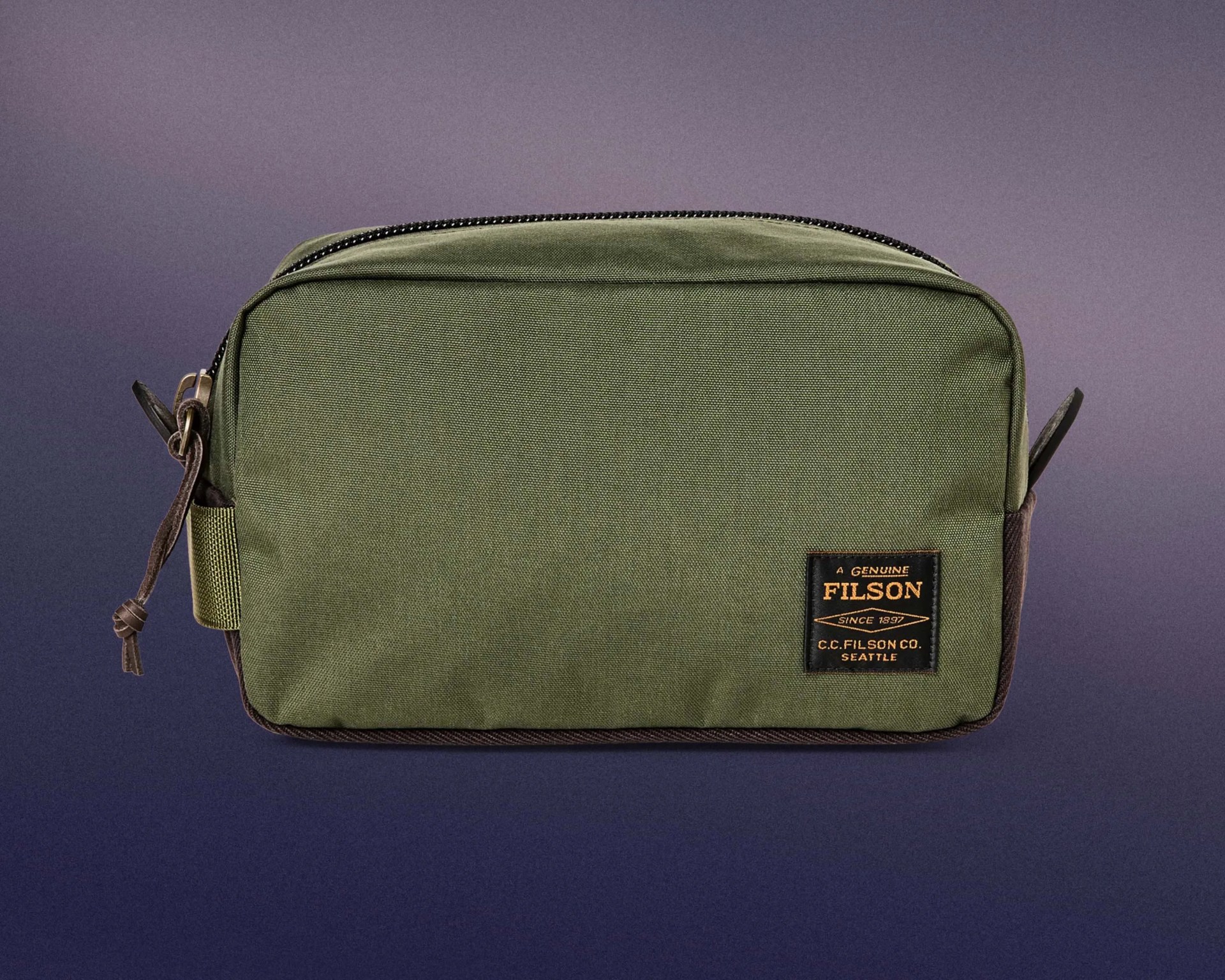 Olive green rectangular toiletry bag with a black and orange Filson brand patch on the front, featuring a zipper closure on top and leather zipper pull. The bag has brown accents on the sides and bottom.