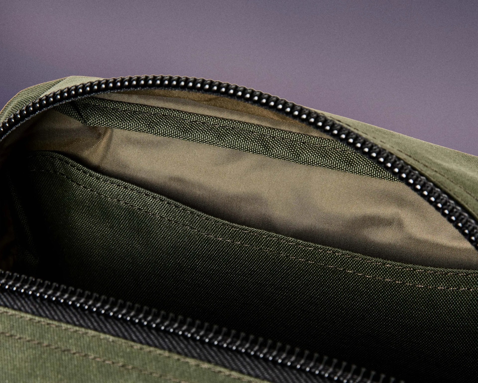 Close-up of the interior of an olive green fabric bag, showing a zippered opening with black teeth and a stitched inner pocket. The lining inside is a lighter olive color.