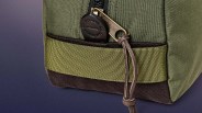 Close-up of a green fabric bag corner with a black zipper pull featuring a leather tab embossed with "FILSON CO. SINCE 1897" and a brass zipper slider engraved with "FILSON." The bag has a textured olive green strap and a dark brown corduroy base, with a brown leather zipper pull string attached. The background is a gradient of dark purple to blue.