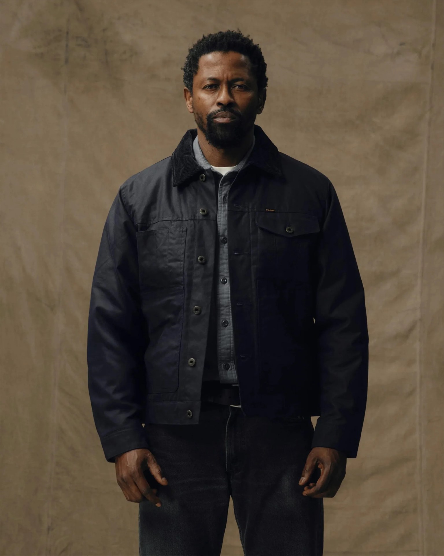 Man wearing a dark navy jacket with a corduroy collar over a gray button-up shirt and black pants, standing against a plain beige background.