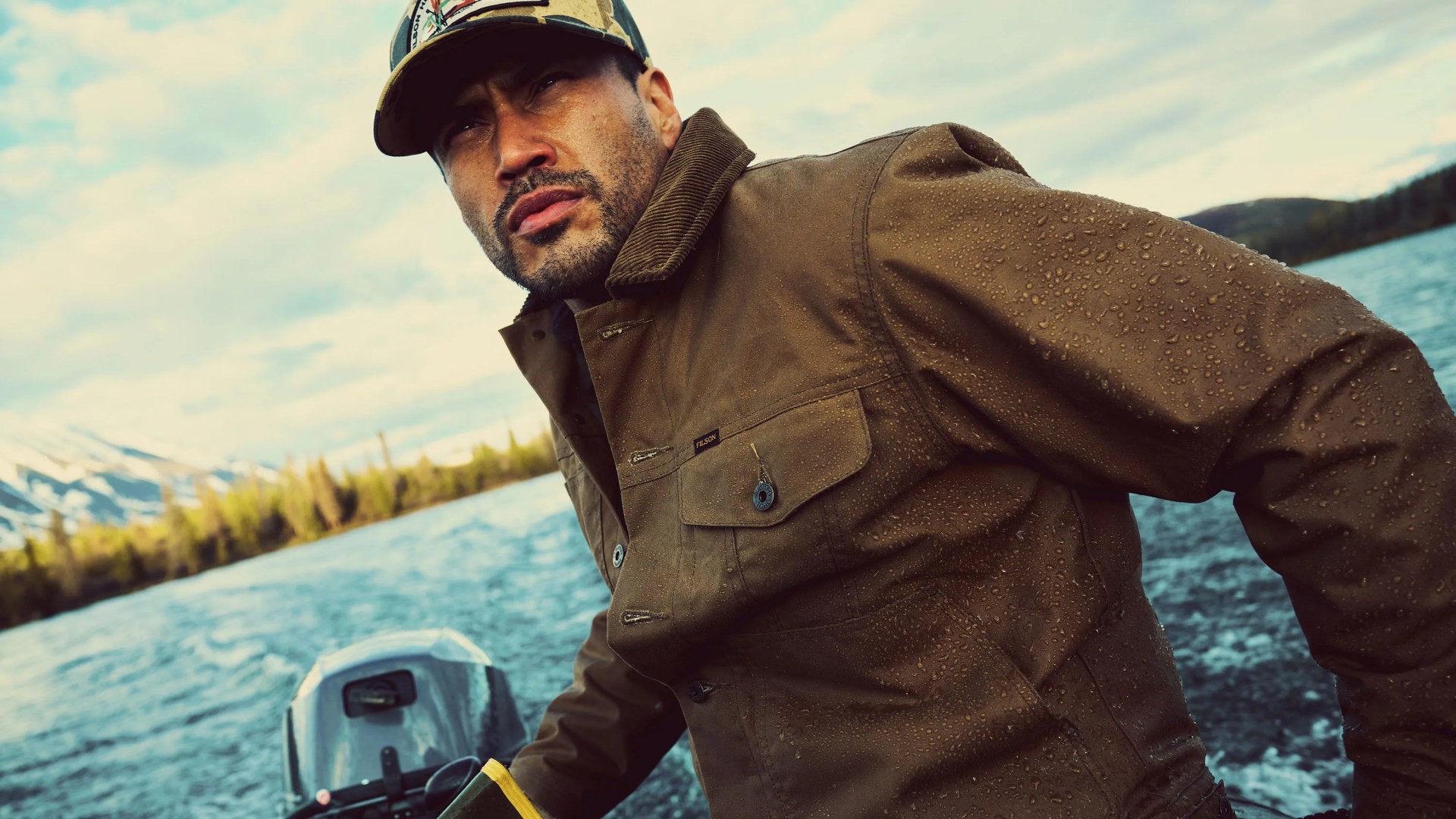 A man wearing a brown water-resistant jacket with visible water droplets and a camouflage baseball cap is steering a boat on a body of water. The background shows a scenic view of trees and snow-capped mountains under a partly cloudy sky.