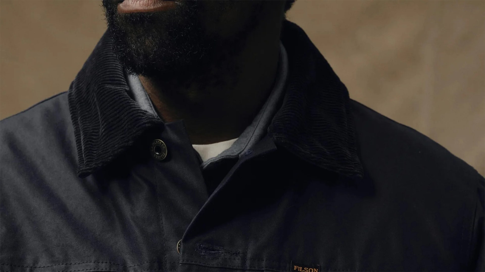 Close-up of a person wearing a dark jacket with a corduroy collar, layered over a gray hoodie and a white shirt underneath. The jacket has a small "FILSON" label on the chest. The person's beard and lower face are partially visible.