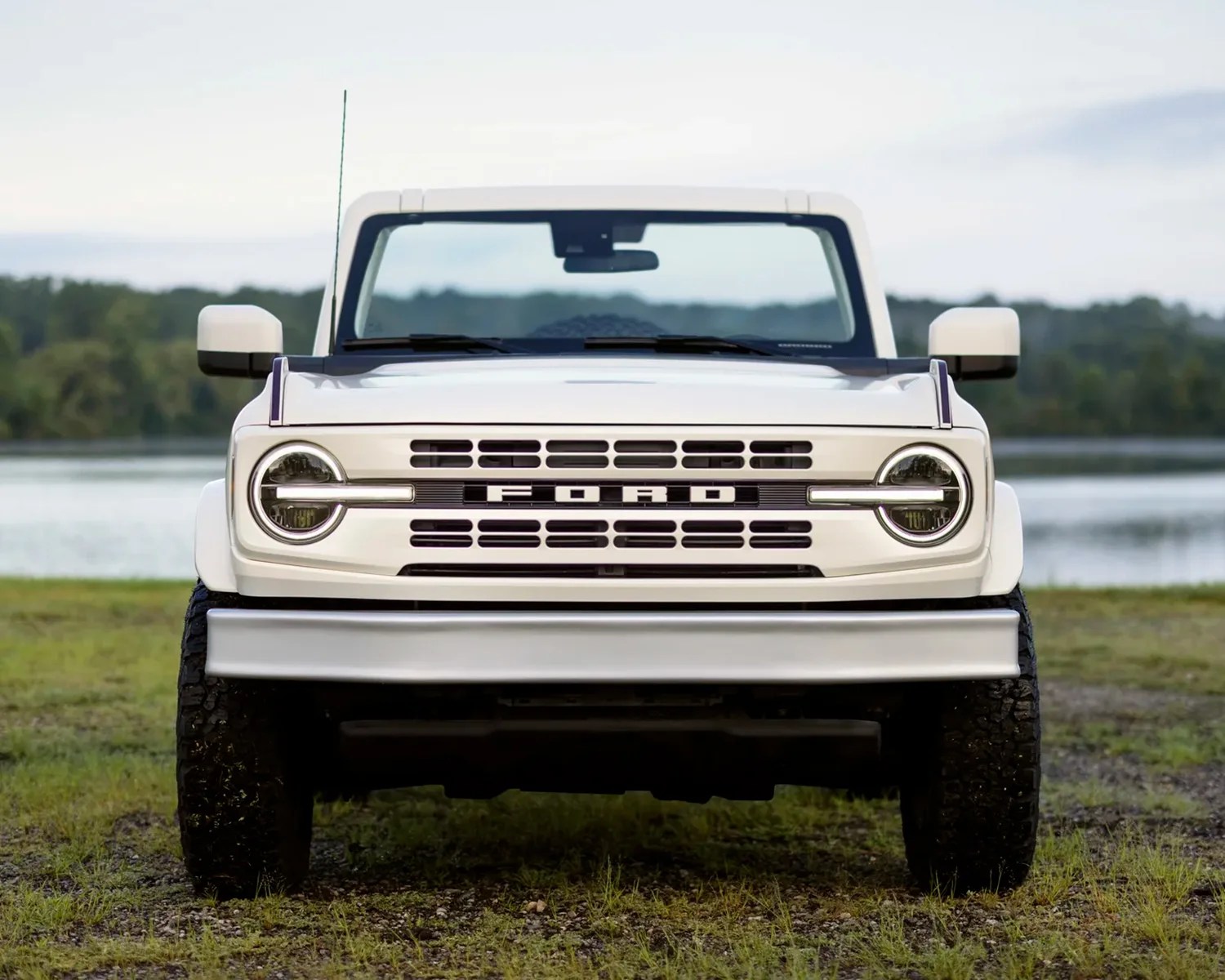 Front view of a white Ford Bronco with round headlights and a bold grille featuring the "FORD" lettering, parked on grass near a body of water with trees in the background.