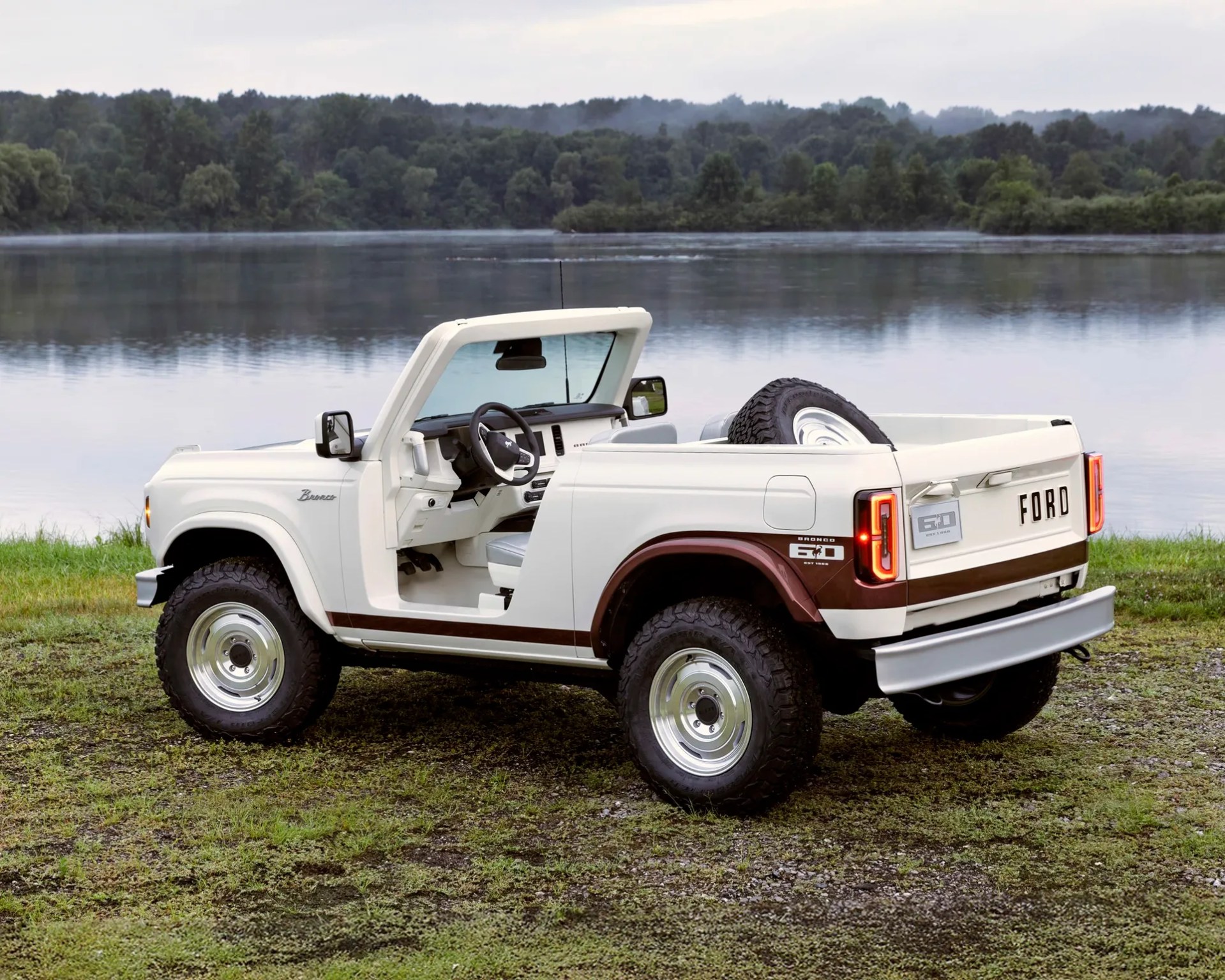 White and brown Ford Bronco 60th Anniversary Edition with no doors, large off-road tires, and a spare tire in the truck bed, parked on grass near a calm lake with a forested background.