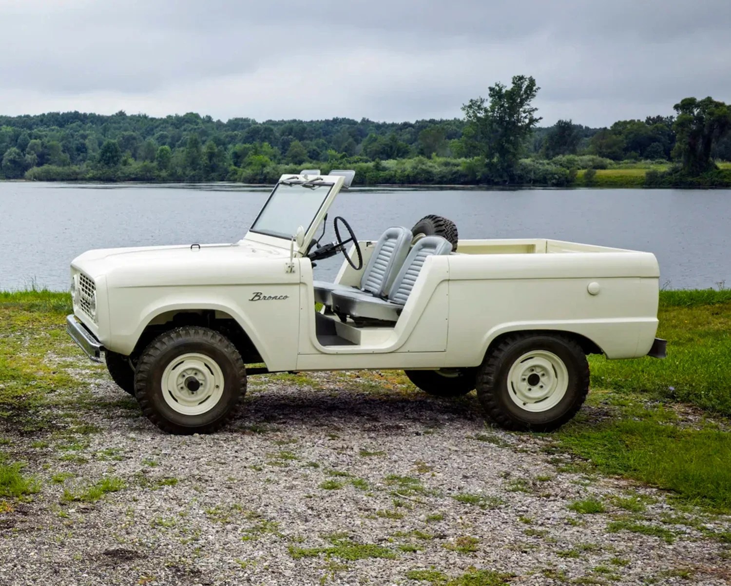 White vintage Ford Bronco with no doors and a convertible top, parked on a gravel area near a body of water with trees in the background.