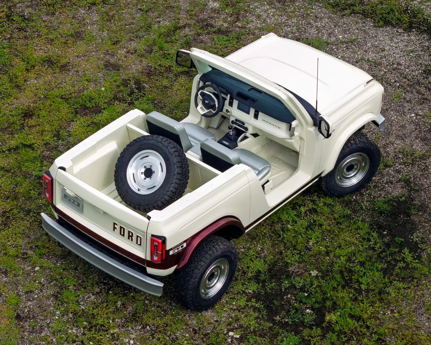 A white Ford Bronco with a maroon stripe around the rear wheel arches, viewed from above. The vehicle has no roof or doors, revealing a simple interior with two front seats and a spare tire mounted in the truck bed. The dashboard features a steering wheel with the Bronco logo and a central display. The vehicle is parked on a patch of grass and gravel.