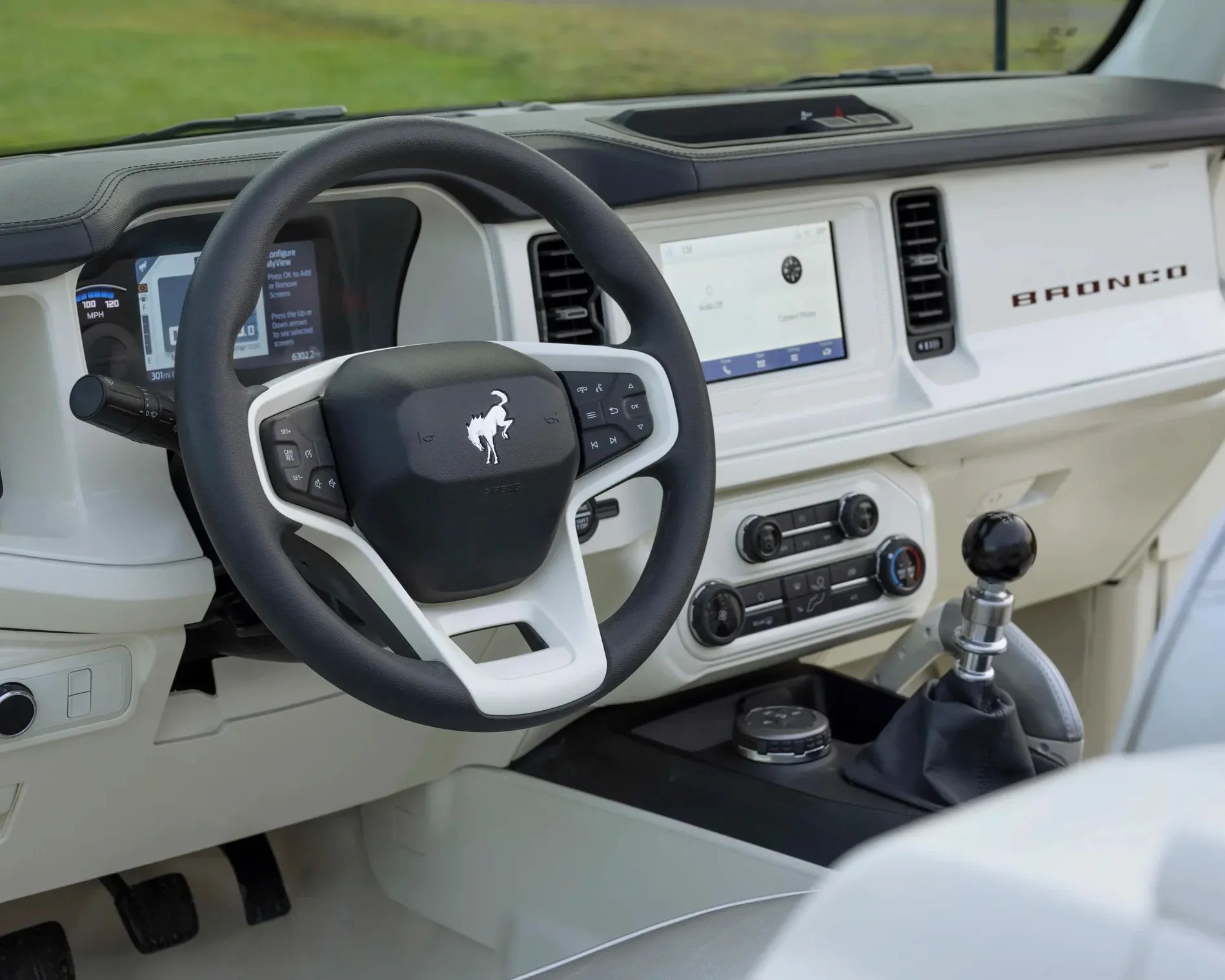 Interior of a Ford Bronco featuring a black and white steering wheel with the Bronco logo, a digital instrument cluster, a touchscreen infotainment system, manual gear shifter with a black knob, climate control dials, and a white dashboard with "BRONCO" lettering.