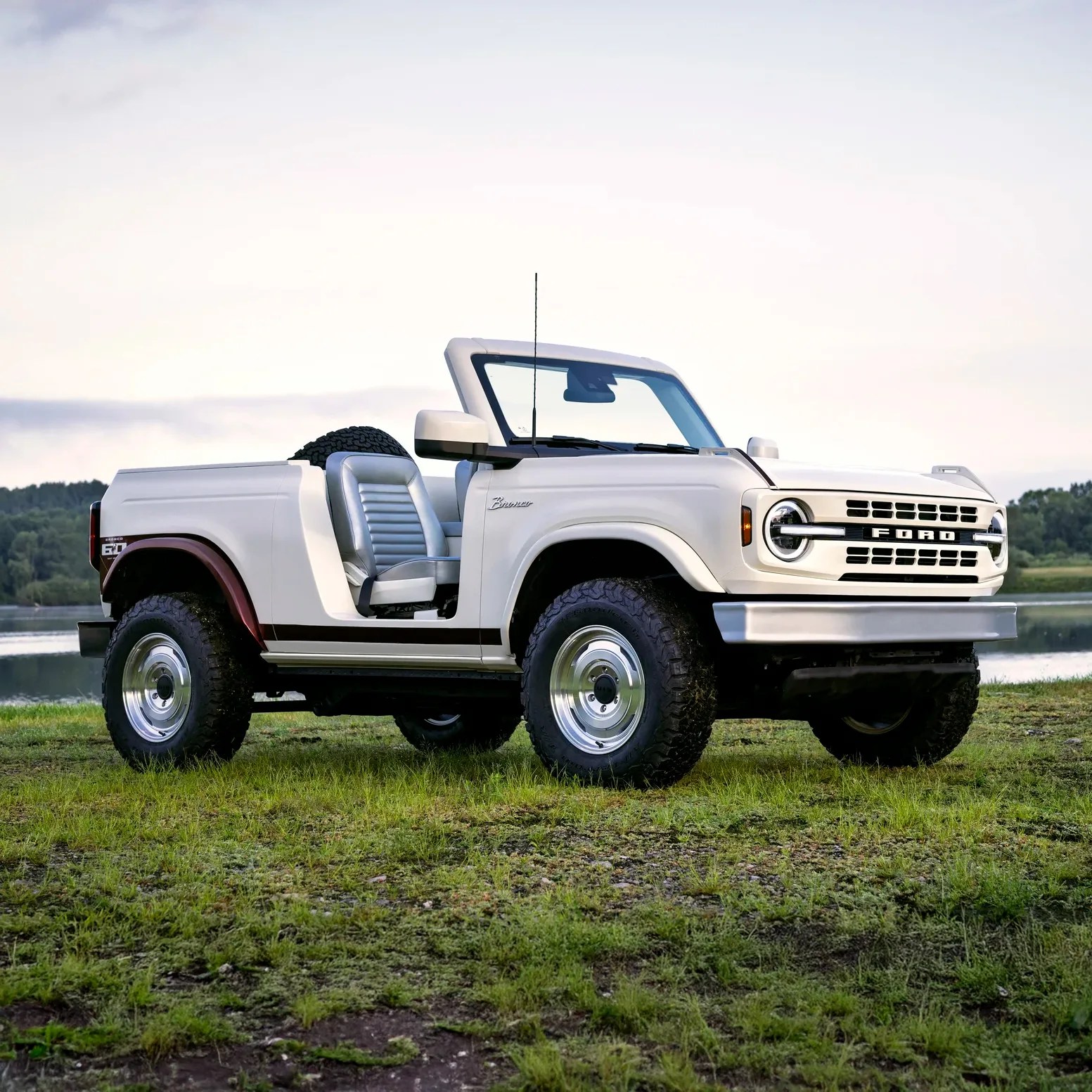 White vintage Ford Bronco convertible with off-road tires, silver seats, and no roof, parked on grassy terrain near a body of water with trees in the background.