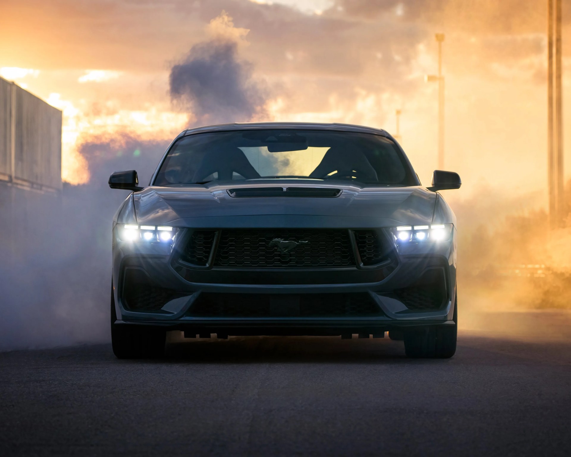 Front view of a dark-colored Ford Mustang with headlights on, surrounded by smoke or mist, set against a sunset or sunrise sky.